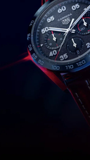 Close-up of luxury men's chronograph watch with black dial and leather strap on dark background.